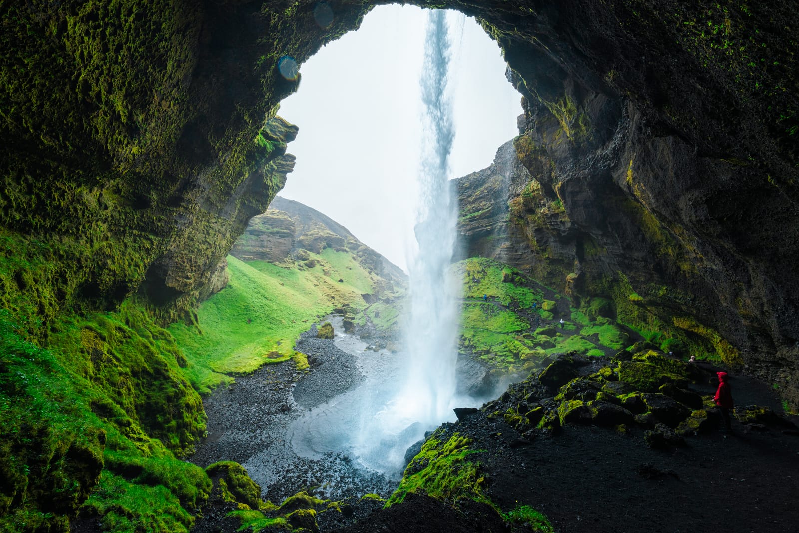 View from inside a mossy cave mouth at a waterfall, Iceland, figure in a red jacket on the path.